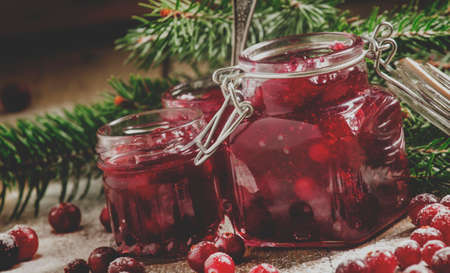 Winter cranberry sauce in glass jars with fresh cranberries, decorated fir branches and snow on the old wooden background, selective focus and toned imageの写真素材