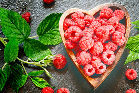 Fresh raspberries with leaves in a bowl in the shape of a heart on a dark background, top view,...
