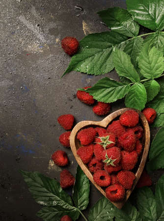 Fresh raspberries with leaves in wooden bowl in the shape of heart on dark kitchen table background,...