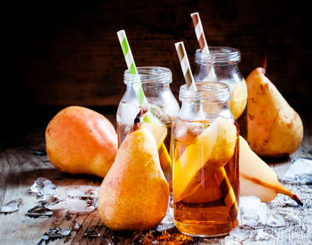 Clarified pear juice with ice and slices of fruit in glass bottles with straws on a dark wooden background, selective focusの写真素材