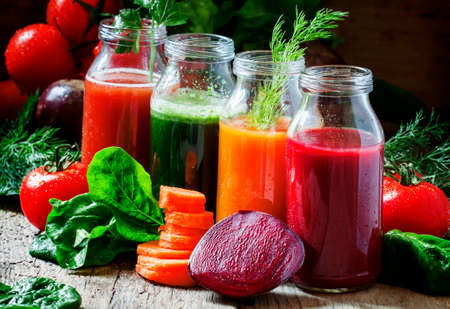 Four kind of vegetable juices: red, burgundy, orange, green, in small glass bottles, fresh vegetables and herbs, vintage wooden background, selective focusの写真素材
