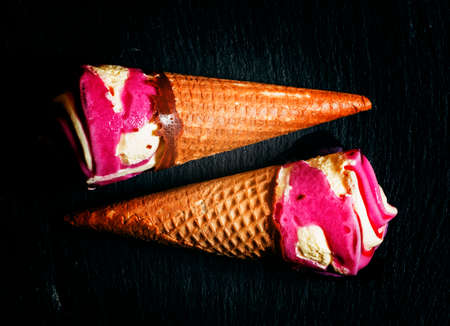 Strawberry and vanilla ice cream in a waffle cone, two on a dark background, top view