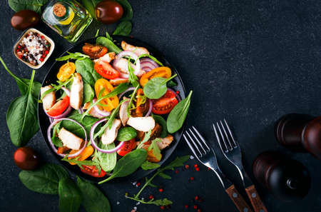Spring salad with spinach, grilled chicken, cherry tomatoes, arugula, corn salad and red onion. Healthy food concept. Black stone table. Top view. Copy spaceの写真素材