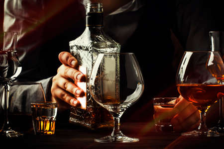The bartender pours the cognac or brandy in big wine glass on the old bar counter. Vintage wooden background in pub or bar, night mood. Place for text, toning, selective focusの写真素材