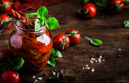 Italian Sun Dried tomatoes in olive oil with green basil and spices in glass jar on wooden kitchen table, rustic style, copy spaceの写真素材