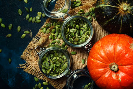 Healthy food, raw pumpkin seeds in glass jars and fresh pumpkins, dark background, top viewの写真素材