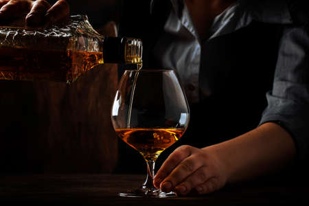 The bartender pours the cognac or brandy in big wine glass on the old bar counter. Vintage wooden background in pub or bar, night mood. Place for text, toning, selective focusの写真素材
