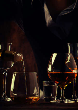 The bartender pours the golden rum in big glass on the old bar counter. Vintage wooden background in...