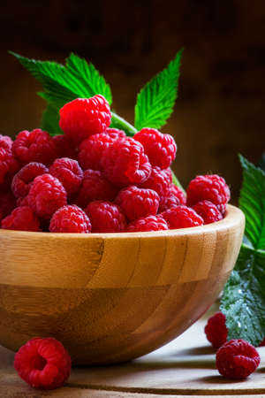 Fresh organic raspberries in bowl, green leaves, vintage wooden background, selective focus