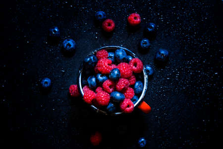 Raspberries and blueberries in old enamel mugs, dark food background, top viewの写真素材