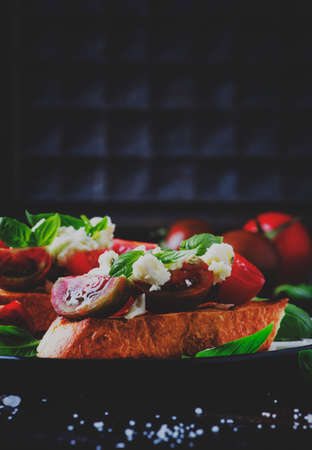 Crispy bruschetta with red and brown cherry tomatoes, feta cheese and green basil on the plate,...