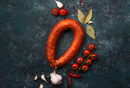 Smoked sausage on gray kitchen table background with aromatic herbs and spices, tomatoes and garlic,...