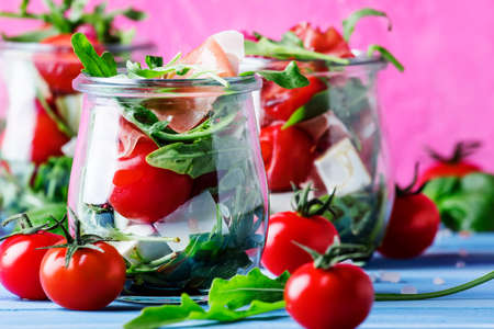 Salad with arugula, cheese, cherry tomatoes and prosciutto in glass jars, pink blue background, selective focusの写真素材