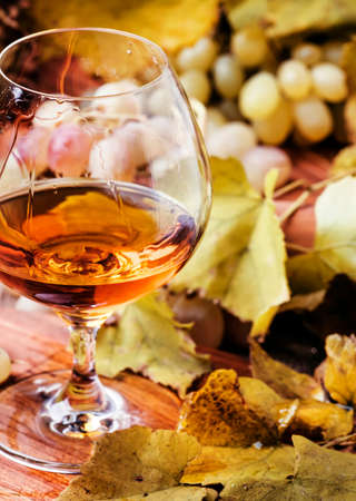 Brandy in a glass and fresh grapes with leaves and a vine, still life in rustic style, vintage wooden background, selective focusの写真素材