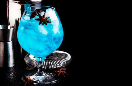 Blue alcoholic cocktail with sambuca, liqueur, lemon juice, crushed ice and anise stars in big wine glass with bartender steel tools, black bar counter background, selective focusの写真素材