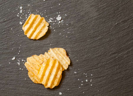 Corrugated potato chips with sea salt on black stone background, top view