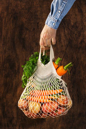 Mesh bag with vegetables and herbs in female hand. Woman hold string net shopping bag on wooden background. Modern reusable shopping, eco friendly lifestyle, zero waste conceptの写真素材