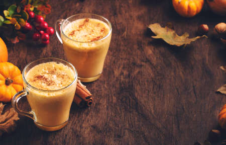 Pumpkin spiced latte or coffee in glass cup on vintage wooden table. Autumn or winter hot drinks and beverages. Fall decor table setting with fallen leaves, small pumpkins, pine cones and nutsの写真素材