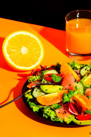 Healthy breakfast with salmon salad with avocado, sesame seeds, tomatoes mixed herbs and orange juice. Contemporary still life, orange black background, hard light and harsh shadowsの写真素材