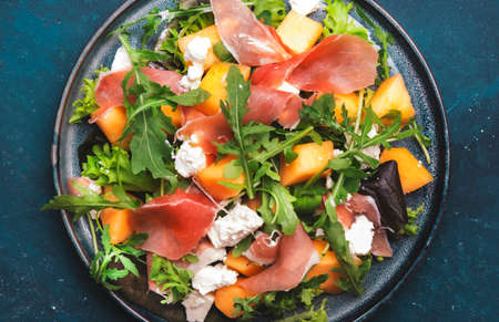 Fresh salad with cantaloupe melon, prosciutto, soft cheese and arugula on blue background, top view,...