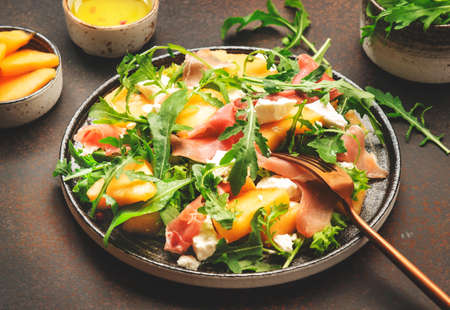 Salad with melon, jamon, goat cheese and arugula on brown background, top view, copy spaceの写真素材