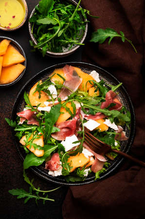 Summer salad with melon, jamon, white cheese and arugula on brown table background, top view, copy spaceの写真素材