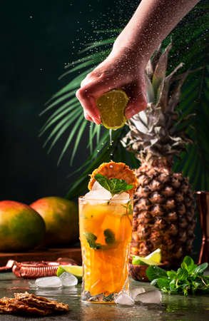 Summer cocktail with vodka, pineapple juice, mango, ice. Long drink or cold mocktail. Bartender hand squeezes lime juice, frozen motion and flying drops. Tropical background with palm leavesの写真素材