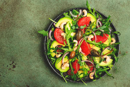 Healthy vegan vegetarian lunch salad with arugula, avocado, grapefruit, cashews, onions, capers and dressing of olive oil, honey and wine vinegar. Green rustic kitchen table, top viewの写真素材