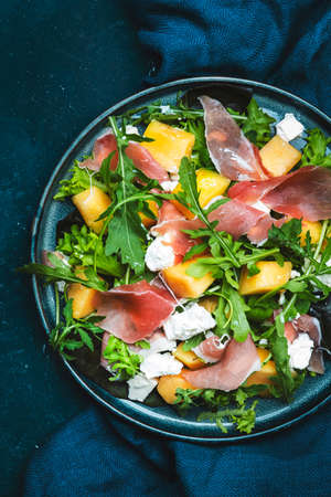 Delicious summer fresh salad with cantaloupe melon, prosciutto, soft cheese and arugula on blue table background, top view, copy spaceの写真素材