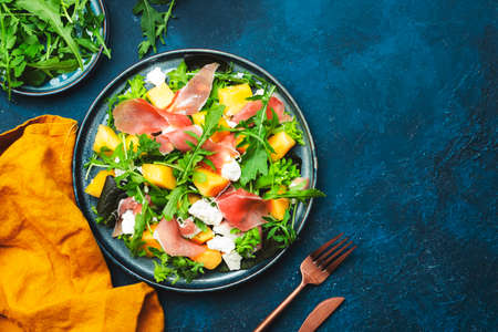 Delicious summer fresh salad with cantaloupe melon, prosciutto, soft cheese and arugula on blue table background, top view, copy spaceの写真素材