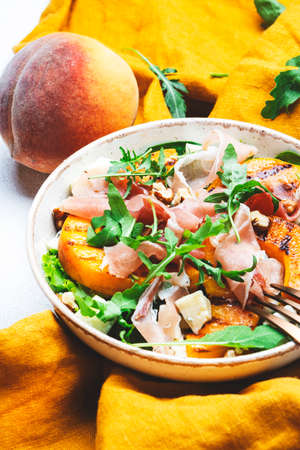 Summer salad bowl with sweet grilled peach, jamon, soft cheese, walnuts and fresh arugula on white kitchen table background, top view, negative spaceの写真素材