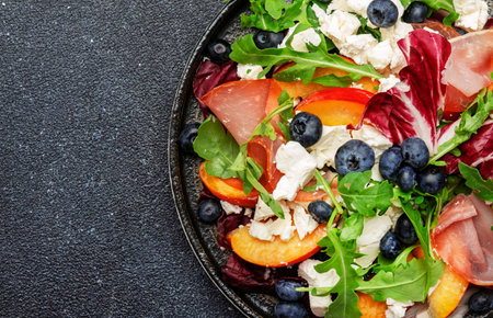 Fresh salad with arugula, radicchio, sweet peaches, smoked pork ham, feta cheese and blueberries. Black stone table background, top view, close upの写真素材