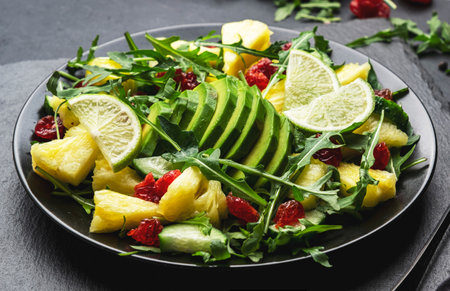 Fresh vegan salad with pineapple, arugula, avocado and dried cranberry on black stone kitchen table,...