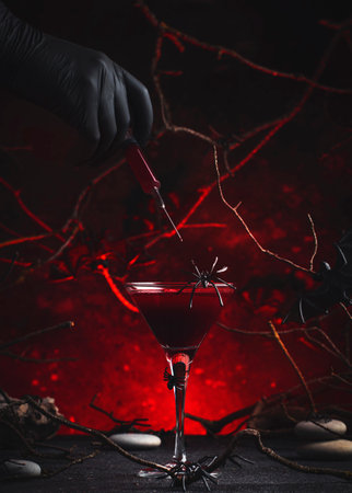 Halloween alcoholic cocktail bloody martini and hand with syringe on scary dark red background with twisted branches, bats, stones, pumpkin guards and spiders, festive drink for vampire partyの写真素材