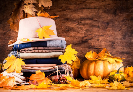 Cozy autumn background with warm autumn and winter clothes, scarves, sweaters, felt hat, gloves, pumpkins, fallen leaves on rustic wood backgroundの写真素材