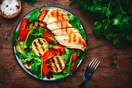 Grilled chicken breast fillet and vegetables salad with paprika, zucchini, eggplant, fresh tomatoes and mixed herbs. Rustic wooden table background, top viewの写真素材