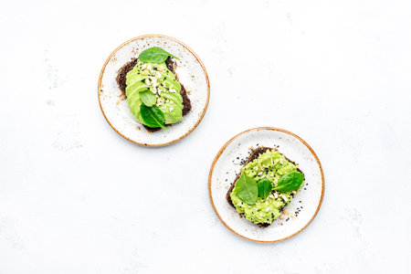 Avocado sandwich or toast on rye bread with spinach, crushed cashew nuts and sesame seeds, on plate, white table background, top viewの写真素材