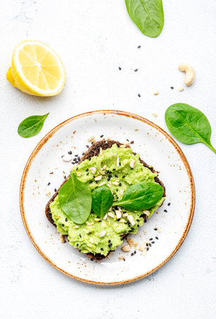 Avocado sandwich or toast on rye bread with spinach, crushed cashew nuts and sesame seeds, on plate, white table background, top viewの写真素材