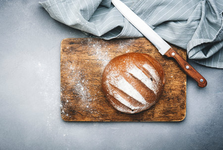 Rye sourdough bread, whole loaf on wooden cutting board, bread knife and kitchen linen towel. Gray...