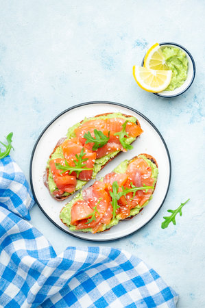 Avocado and salmon sandwich on rye bread with guacamole sauce, arugula and sesame seeds on plate, blue table background, top viewの写真素材