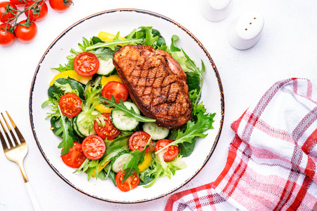 Fresh salad with grilled duck breast with red tomato, cucumber, paprika and chard, frisse, mizuna and arugula leaves on white table background, top viewの写真素材