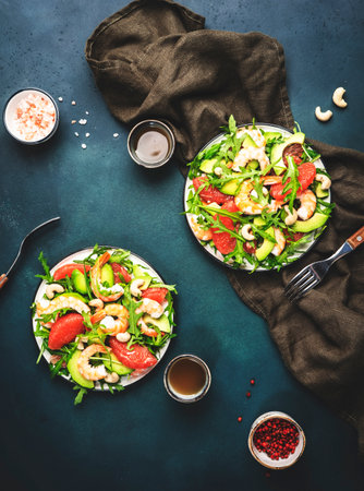 Two with salad with shrimps, avocado, grapefruit plates, arugula and cashews, table setting. Dark green background, top viewの写真素材