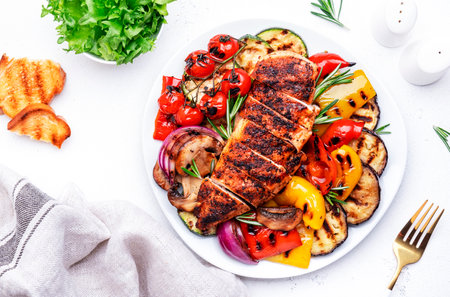 Grilled vegetables and chicken breast fillet. Colorful paprika, zucchini, eggplant, mushrooms, tomatoes, onion and rosemary, served on plate, white table background, top viewの写真素材