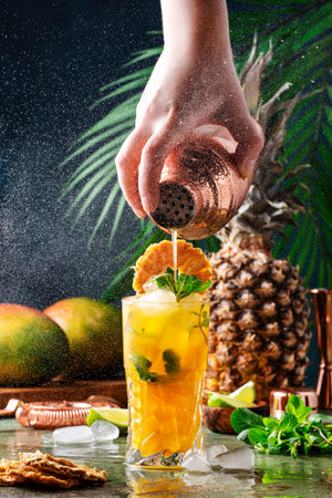 Summer cocktail with vodka, pineapple juice, mango, ice. Long drink or cold mocktail. Bartender pours drink from copper shaker, frozen motion and flying drops. Tropical background with palm leavesの写真素材