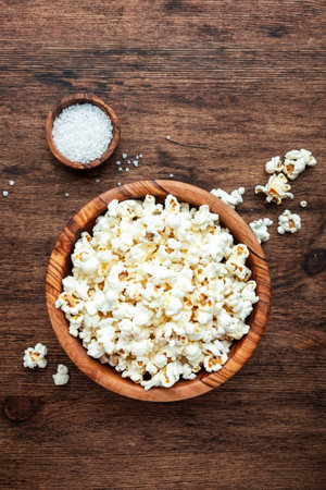 Salted popcorn in wooden bowl, unhealthy food, wooden kitchen table background, top viewの写真素材