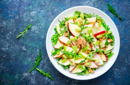 Waldorf salad with green and red apple, celery, lettuce, chicken fillet, arugula and crunchy walnuts on plate, blue table background, top viewの写真素材