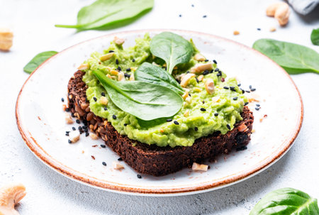 Avocado toast on rye bread with spinach leaves, cashew nuts and sesame seeds on plate, white table...