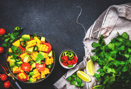 Vegan salad with sweet corn, avocado, jalapeno pepper, red tomatoes, fresh cilantro and olive oil and lime dressing. Black table background, top viewの写真素材