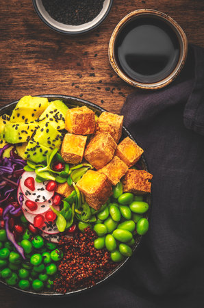 Vegan buddha bowl with red quinoa, fried tofu, avocado, edamame beans, green peas, radish, red cabbage and sesame seeds with soy sauce. Healthy diet food. Wood kitchen table background, top viewの写真素材