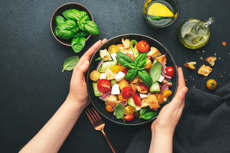 Hands holding plate with panzanella salad with stale ciabatta bread, colorful tomatoes, soft cheese, red onion, olive oil, sea salt and green basil, black table background, top viewの写真素材